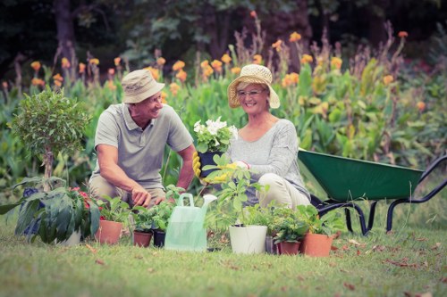 A smiling elderly couple wearing sun hats is working together in a well-maintained garden near Crystal Palace, surrounded by flowering plants, shrubs, and potted greenery. They are seated on the grass, with garden tools and plant pots arranged around them, and a green wheelbarrow is visible nearby. In the background, there is a lush border of purple and orange flowers along with leafy green plants, indicating a vibrant and cultivated outdoor space typical of a residential garden in the Crystal Palace area. The scene is bathed in natural daylight, suggesting a clear, pleasant day suitable for gardening or outdoor maintenance services provided by Landscapers Crystal Palace, with a focus on lawn and garden care in the local community.