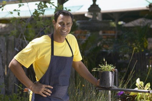A smiling man wearing a yellow T-shirt and a dark apron stands in a well-maintained garden, holding a black plastic pot with a newly planted green shrub. The garden features a neatly trimmed lawn in the foreground, with a variety of plants and tall grasses around the edges. In the background, there are trees, shrubs, and a garden feature such as a fountain or birdbath, along with a wooden trellis partially covered with climbing plants. The sunlight indicates clear weather, casting natural light across the garden scene. The setting appears to be an outdoor space in Crystal Palace, and reflects well-organized landscaping and gardening work typical of local garden maintenance services provided by Landscapers Crystal Palace. The image emphasizes outdoor gardening care, planting activities, and garden enhancement efforts, aligning with services related to lawn care, planting, and outdoor space renovation.