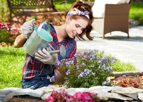 A young woman with light skin, brown hair tied back with a patterned headband, wearing a checkered shirt and gardening gloves, is kneeling in a well-maintained front garden in Crystal Palace. She is smiling as she waters a flowering plant with light purple blooms using a metal watering can. The garden features a neatly edged flower bed with vibrant pink, purple, and green foliage, surrounded by a low stone border. In the background, there are paving slabs and a portion of a garden patio area with outdoor furniture, including a single chair. The scene takes place under natural daylight with clear weather, highlighting the lush greenery, healthy soil, and the colorful, thriving plants indicative of professional gardening services offered by Landscapers Crystal Palace. The environment appears tidy and well-kept, reflecting ongoing outdoor maintenance and garden care in a residential setting.