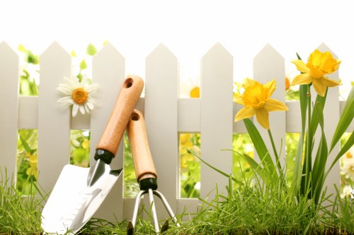 A close-up view of a small garden border in a residential outdoor space, featuring a white wooden picket fence in the background. In the foreground, there is lush green grass with a mixed flower bed containing vibrant yellow daffodils and white daisies growing alongside the fence. A pair of gardening tools—a trowel and a hand cultivator with wooden handles—are leaning against the grass, indicating recent planting or garden maintenance. The soil within the flower bed appears freshly turned, and the garden is well-lit by natural sunlight, suggesting a clear, bright day. The scene captures the neat arrangement of flowering plants and the well-kept lawn, illustrating typical garden features that a gardening professional such as Landscapers Crystal Palace might work on to enhance outdoor spaces in the Crystal Palace area or surrounding postcode district, reflecting the natural tones and seasonal beauty involved in professional gardening and planting services.
