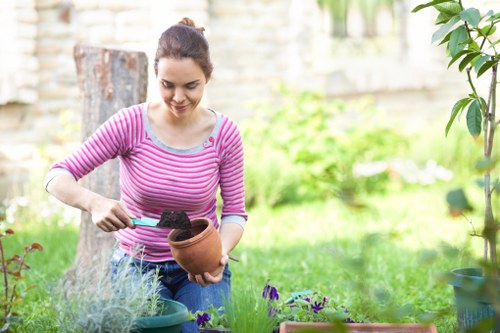 A woman with dark hair tied back, wearing a pink and grey striped long-sleeve shirt, is engaged in gardening in an outdoor backyard space. She is holding a small terracotta pot filled with soil in her left hand while using a trowel in her right hand to scoop soil into the pot. The garden features a lush, green lawn in the foreground, with a flower bed containing various plants and flowers in the midground. Behind her, there is a weathered tree stump and a light-colored wooden fence that encloses the area. Nearby, potted plants in different containers are visible on the ground, and some foliage from a tall plant with broad leaves extends into the right side of the frame. The weather appears clear and bright, suggesting a sunny day suitable for outdoor gardening activities, which aligns with landscaping and garden maintenance services provided by Landscapers Crystal Palace in the area near the postcode of Crystal Palace, London.