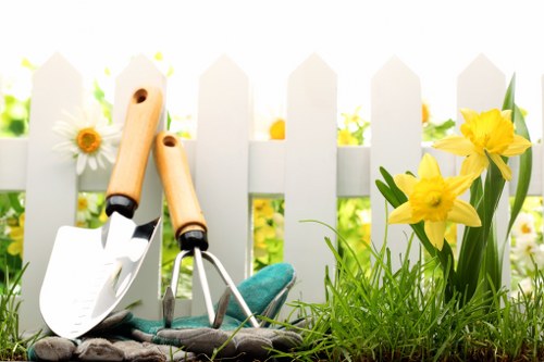 The image depicts a section of a garden with a white picket fence in the background, likely within a residential outdoor space in the Crystal Palace area. In the foreground, there is a patch of lush, green grass with a textured surface, alongside a small flower bed bordered by the fence. The flower bed contains vibrant yellow daffodils with elongated green leaves, adding a splash of colour against the natural tones of the soil and plants. Resting on the grass are gardening tools, including a small hand trowel with a wooden handle and a metal blade, and a transplanting fork with a similar wooden handle, both showing signs of use. The overall scene suggests a tidy, well-maintained garden space suitable for ongoing planting and lawn care activities, characteristic of local garden maintenance services offered by companies like Landscapers Crystal Palace. The weather appears bright and sunny, with natural light illuminating the scene, making the colours vivid and details clear, creating a welcoming outdoor environment for gardening enthusiasts or homeowners seeking garden improvement in the Crystal Palace postcode area.